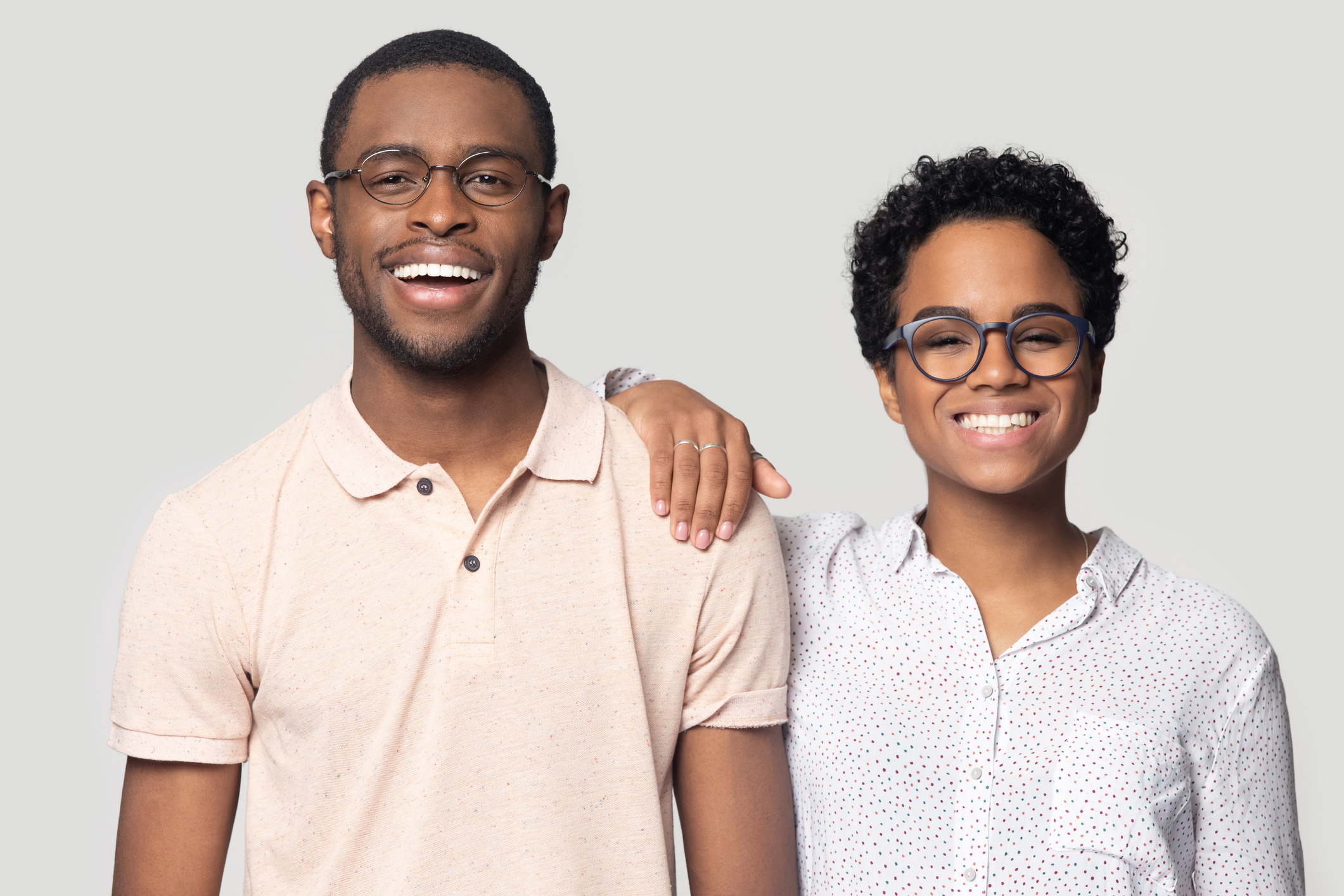 Smiling excited african american married family couple in eyeglasses looking at camera. Young happy black woman leaning on husbands shoulder, posing for photo, isolated on grey studio background.